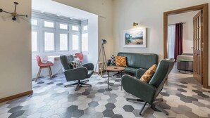 Un salon moderne avec un canapé vert, deux fauteuils, une table basse et un sol orné d’un motif géométrique. Il y a une fenêtre avec des cadres blancs et un tableau accroché au mur.