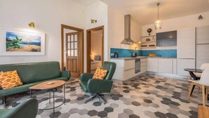 Un salon moderne avec un canapé et un fauteuil verts, une petite table basse et un sol à motifs hexagonaux. On aperçoit, au fond, une cuisine avec des meubles blancs et un crédence bleue.