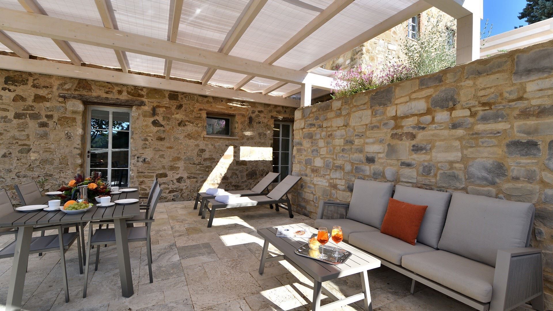 A stone-walled outdoor patio with a covered seating area, a dining table set with plates and a fruit bowl, and a sofa with cushions.
