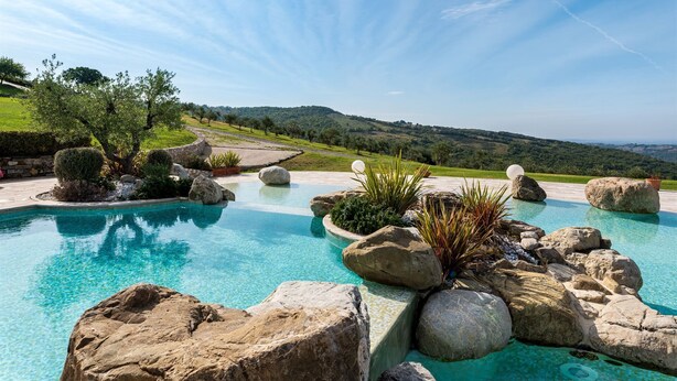 Piscina com água cristalina, rodeada por pedras grandes e plantas, com colinas verdes e céu limpo ao fundo.