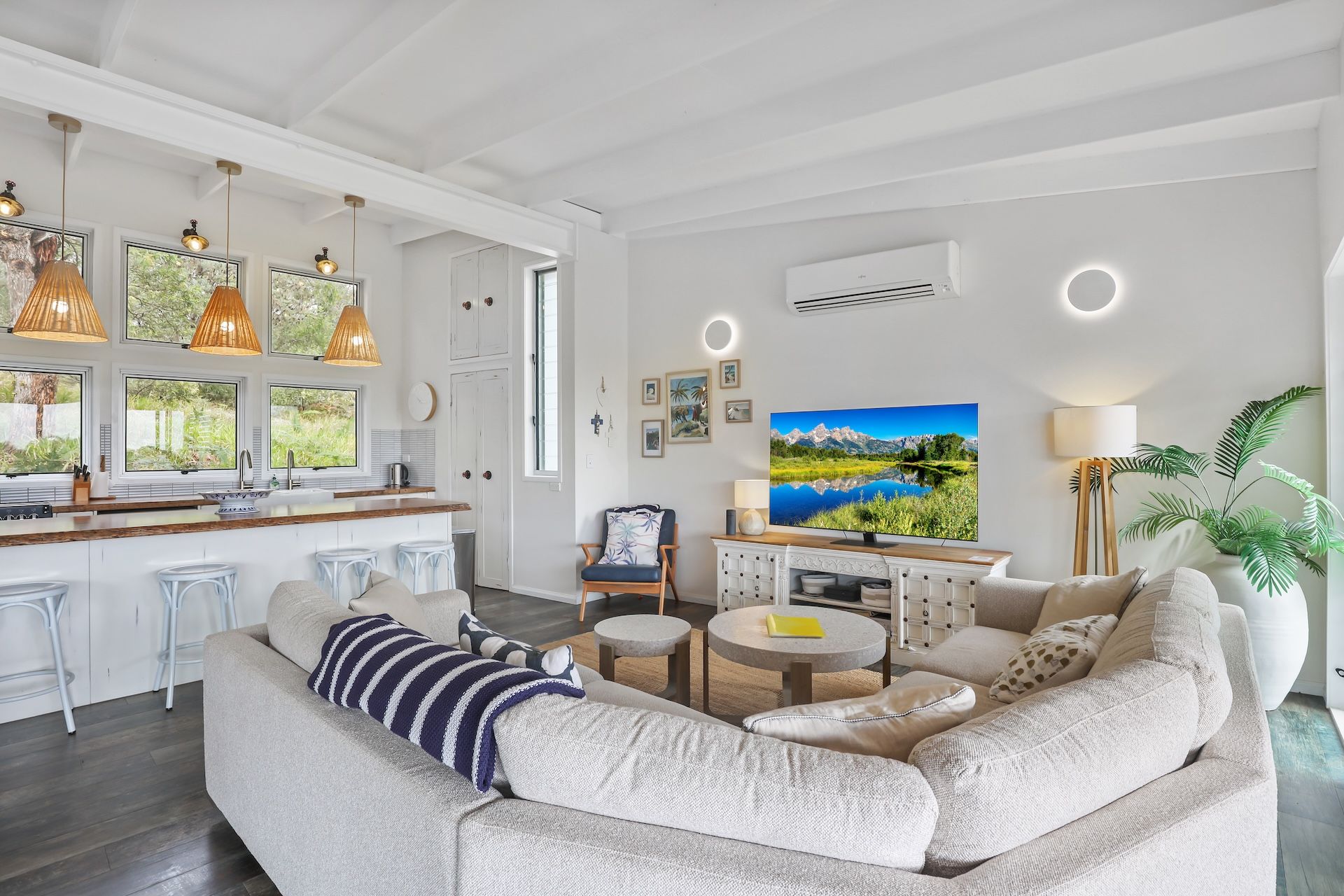 Ein modernes Wohnzimmer mit Sofa, Couchtisch und Fernseher. Es gibt eine Küchenzeile mit Barhocker und Pendelleuchten.