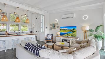Ein modernes Wohnzimmer mit Sofa, Couchtisch und Fernseher. Es gibt eine Küchenzeile mit Barhocker und Pendelleuchten.