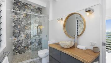 Une salle de bain moderne avec une douche en verre, un meuble-lavabo en bois, un miroir rond et une vue sur la mer.