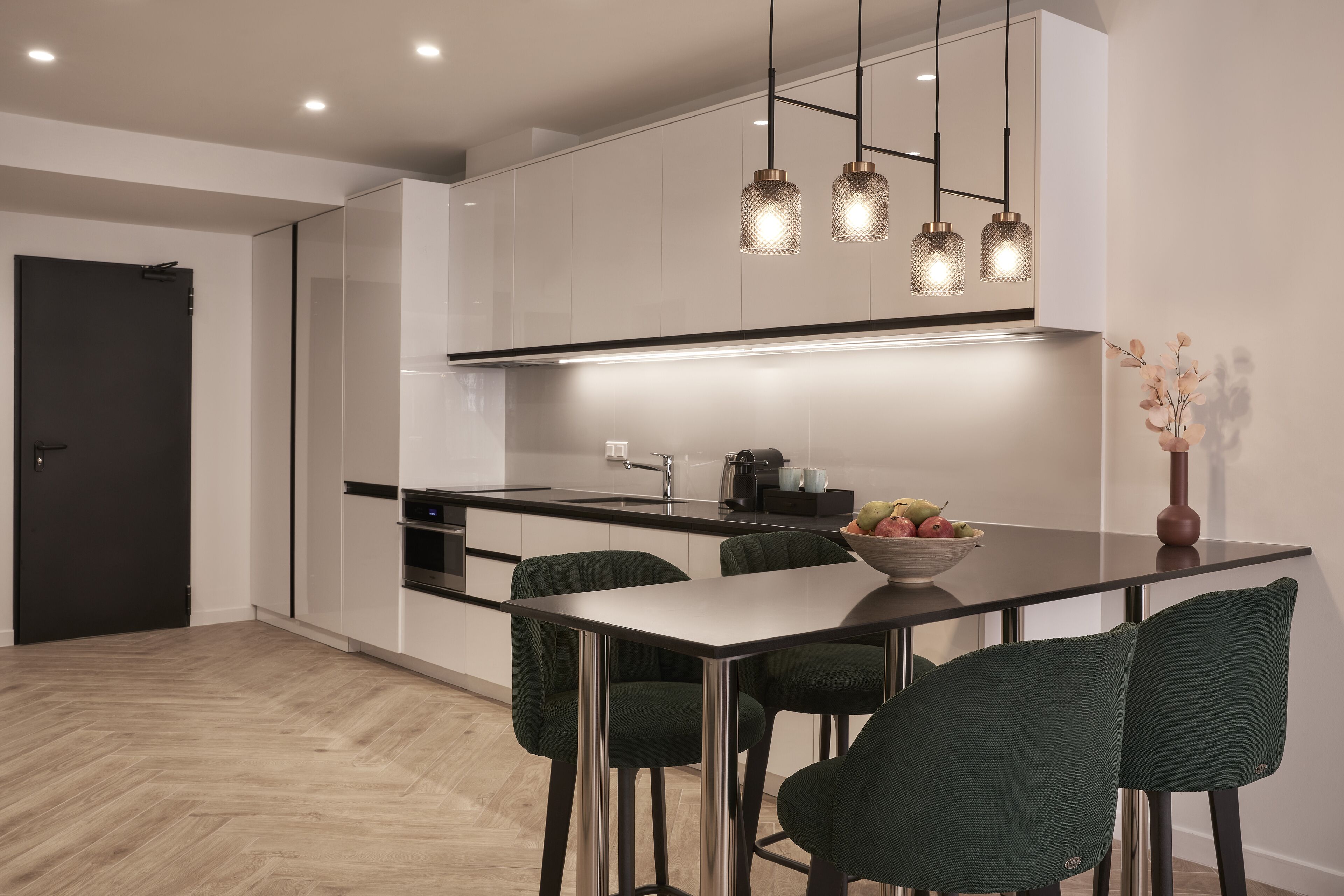 Une cuisine moderne avec des meubles blancs, un plan de travail noir et des chaises vertes. Des luminaires suspendus sont placés au-dessus du plan de travail, et un saladier rempli de fruits se trouve sur la table.