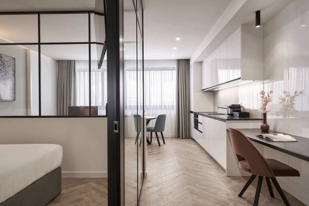 A modern hotel room with a bed, a desk, a chair, a dining table, and a kitchen area.