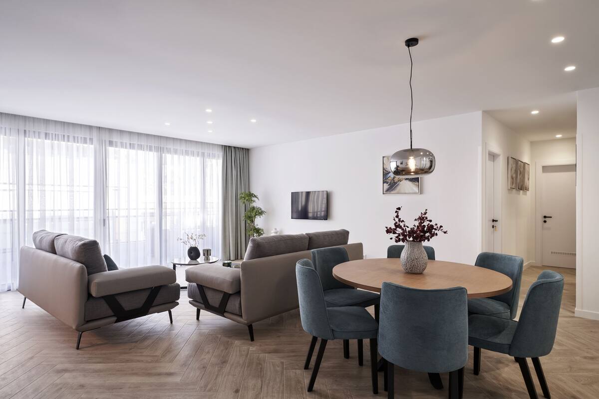 A modern living room with a sofa, armchairs, a dining table with chairs, a TV mounted on the wall, and a large window with curtains.