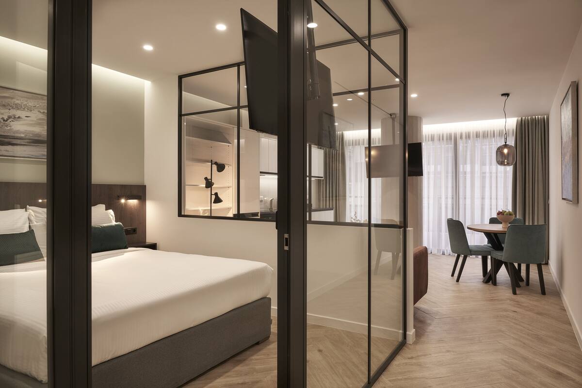 A modern hotel room with a large bed, a dining area with a table and chairs, and a large mirror reflecting the room.