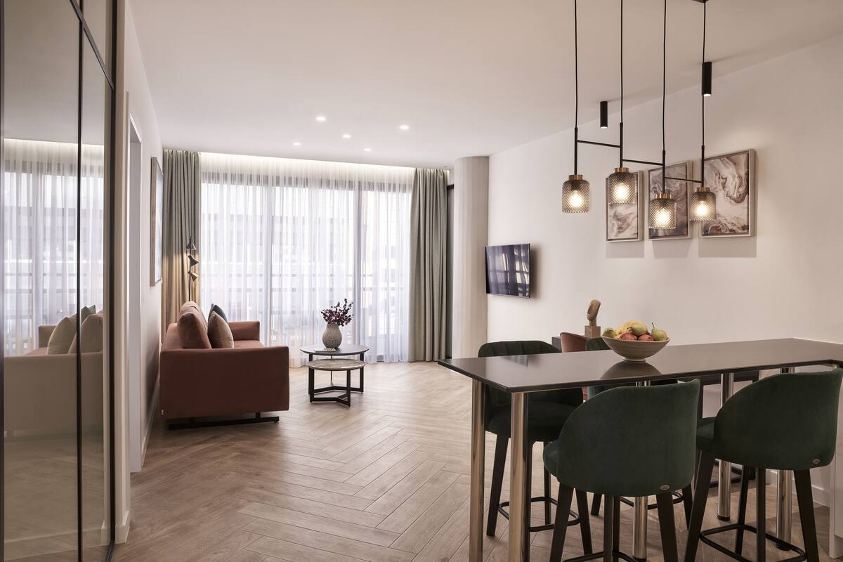 A modern living room with a sofa, a dining table with chairs, and a large window with sheer curtains.