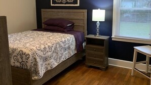 Une chambre à coucher avec un lit en bois, une table de chevet, une lampe, un ventilateur de plafond, un élément décoratif mural et une fenêtre avec des stores.