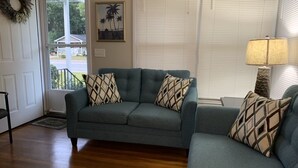 Un salon avec un canapé vert, des coussins à motifs, un plancher en bois, une porte décorée d’une guirlande, une fenêtre avec des stores et une lampe.