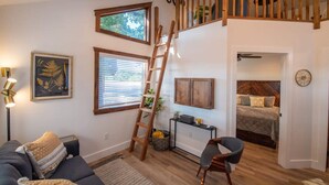 A cozy living room with a wooden staircase, a sofa, a desk, and a bed in the adjacent room.