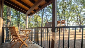 A wooden deck with a chair and table, surrounded by a metal fence, overlooking a wooded area with a small cabin.