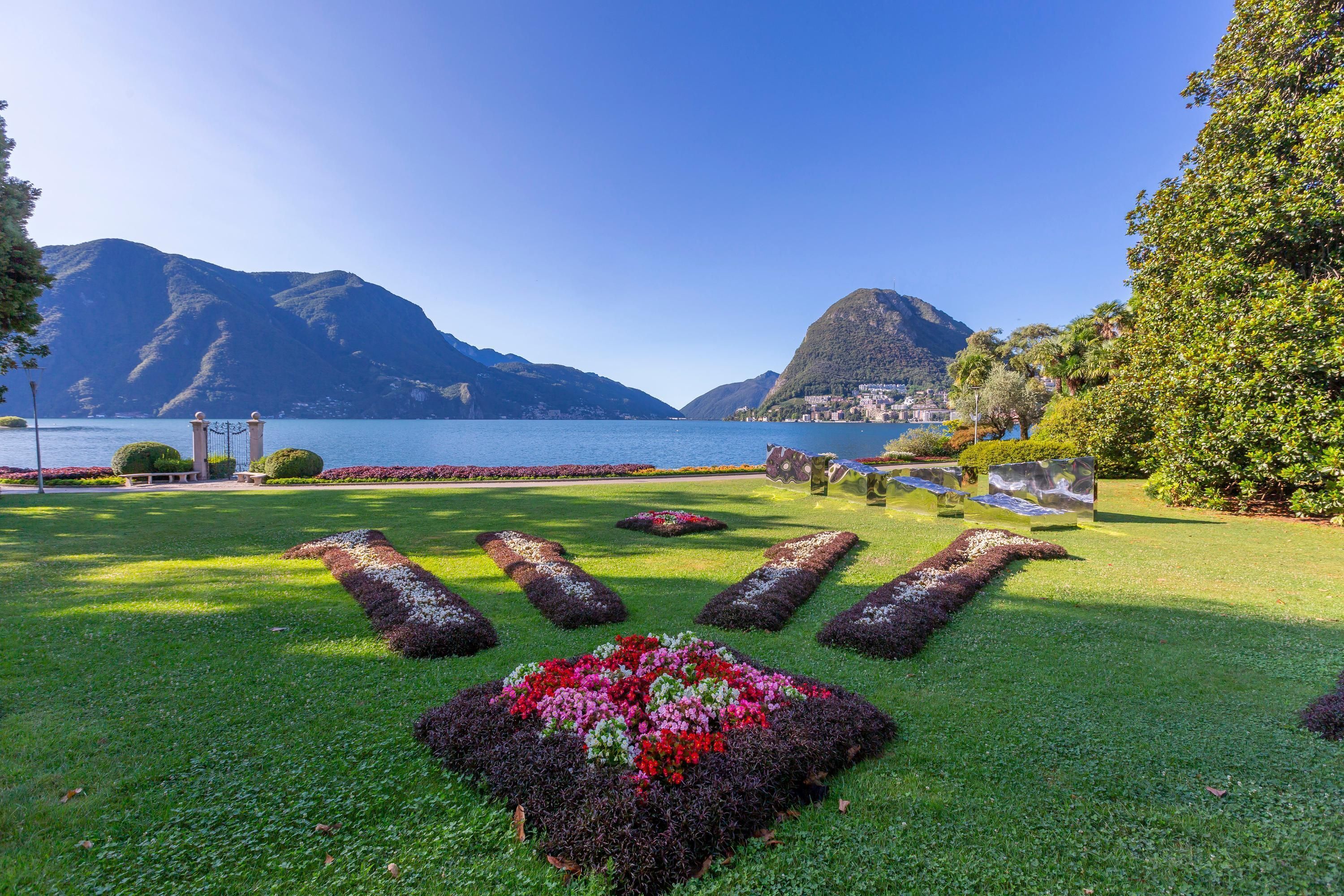 A well-maintained garden with a central flower bed, a pathway leading to a gate, and a view of a lake with mountains in the background.