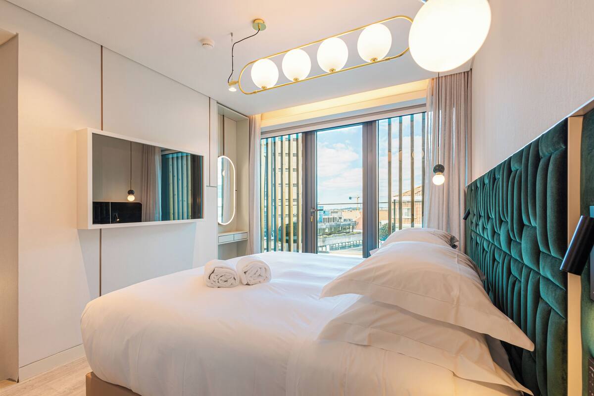 A modern hotel room with a large bed, a built-in TV, and a view of the city through the window.