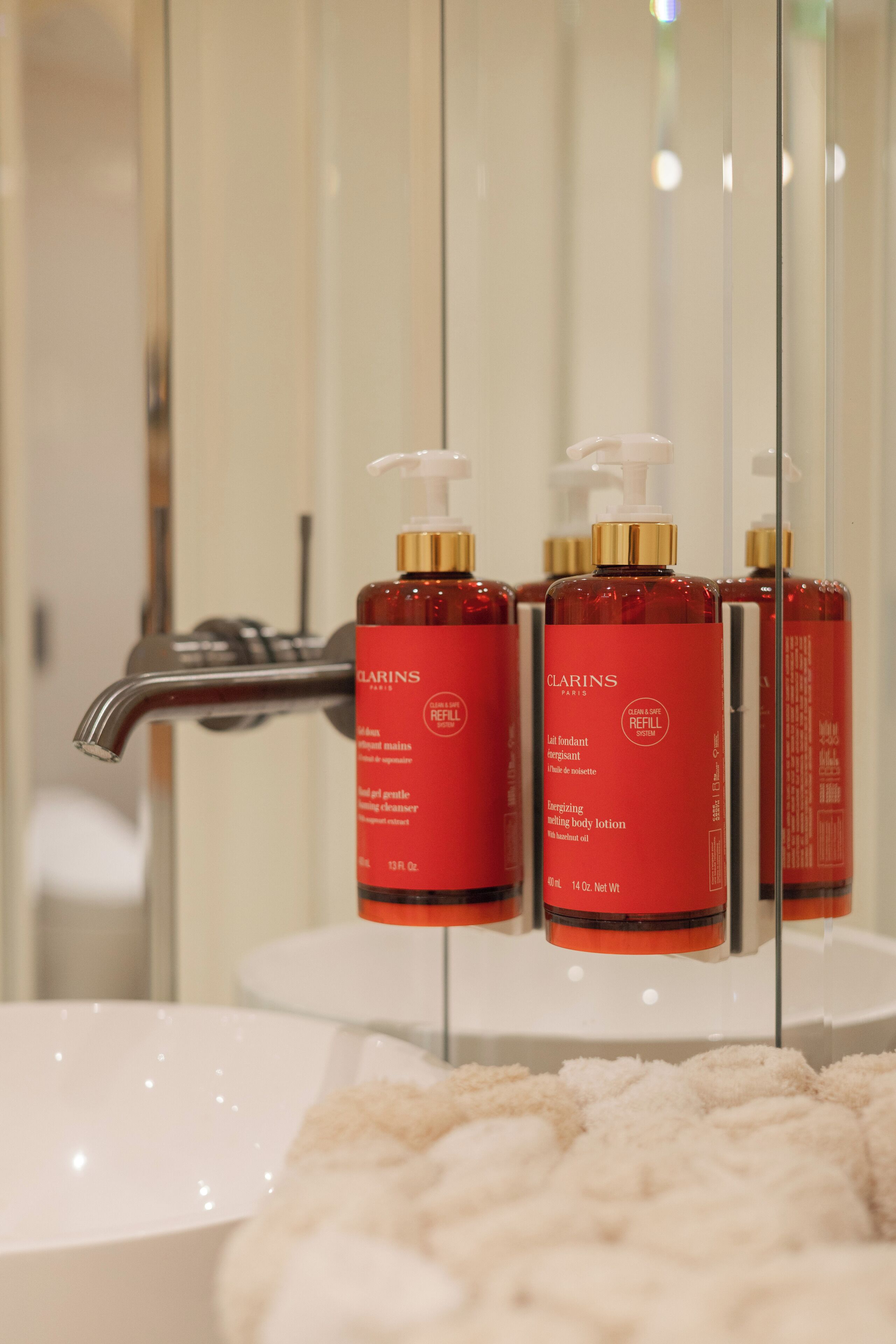 A bathroom with a white sink, a mirror, and three red bottles of Clarins Paris refill system body lotion.