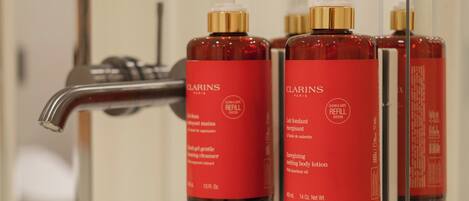 A bathroom with a white sink, a mirror, and three red bottles of Clarins Paris refill system body lotion.