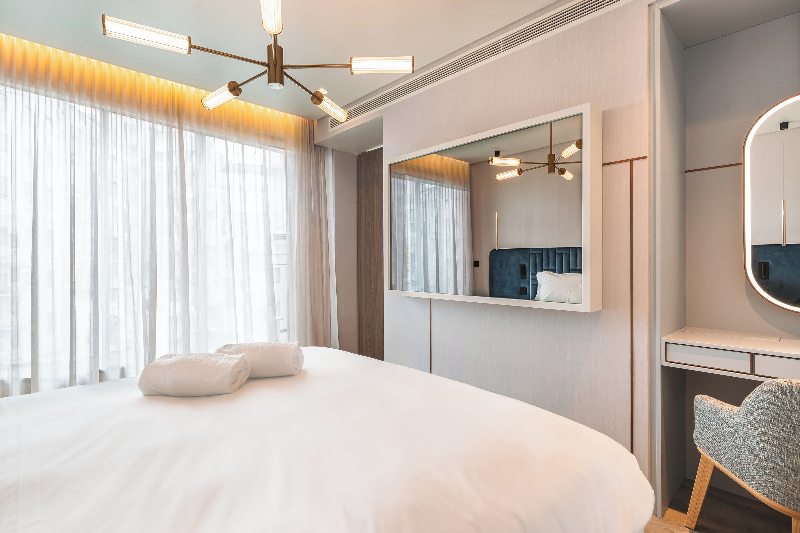 Una habitación de hotel moderna con una cama grande, un escritorio con silla, un espejo amplio y una lámpara en el techo.