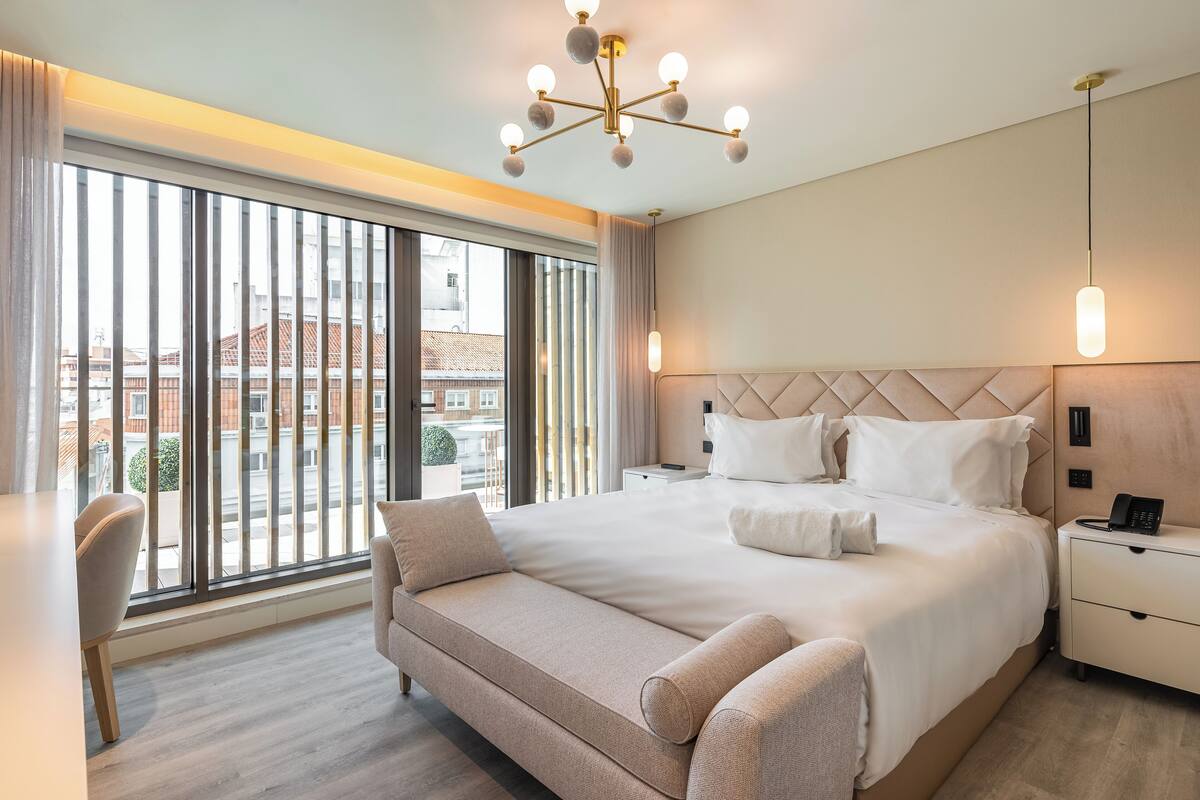 A modern hotel room with a large bed, a sofa, a desk, and a view of the city through floor-to-ceiling windows.