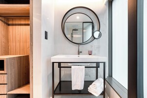 A modern bathroom with a wooden vanity, a round mirror, a white sink, and a glass shelf with towels.