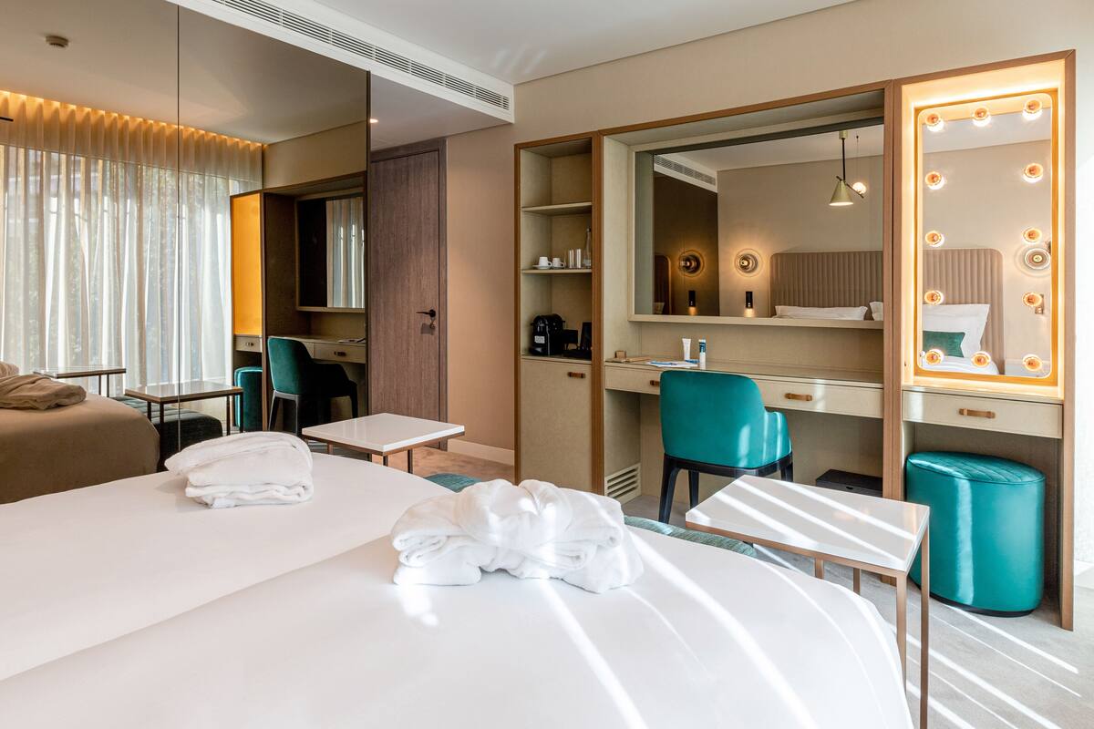 A hotel room with a bed, a desk, a chair, a mirror with lights, a wardrobe, and a bathroom area.