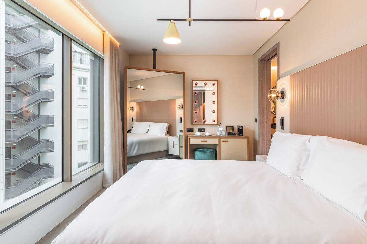A hotel room with a large bed, a desk with a mirror, and a view of a building with balconies.