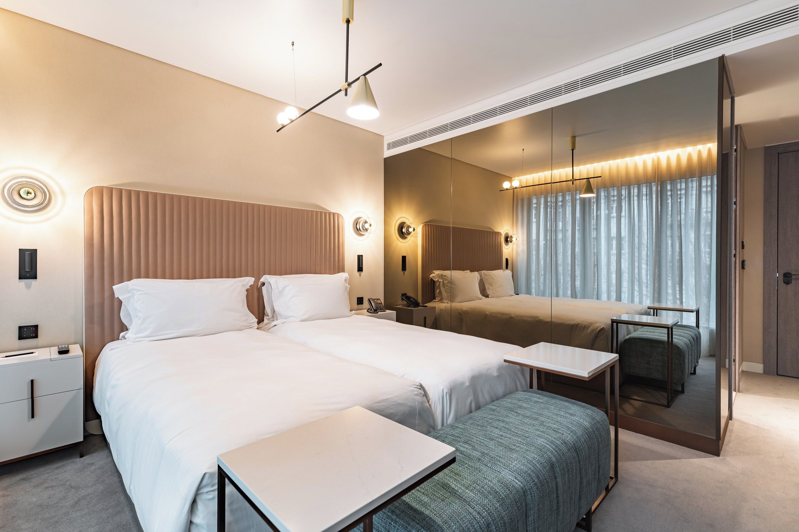 A modern hotel room with two beds, a bedside table, a mirror, and a large window with curtains.