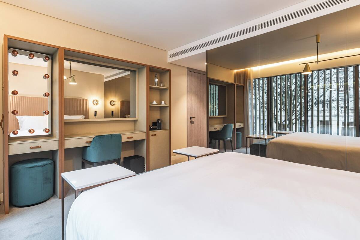 A modern hotel room with a large bed, a desk with a chair, a vanity with a mirror, and a window with a view of trees.
