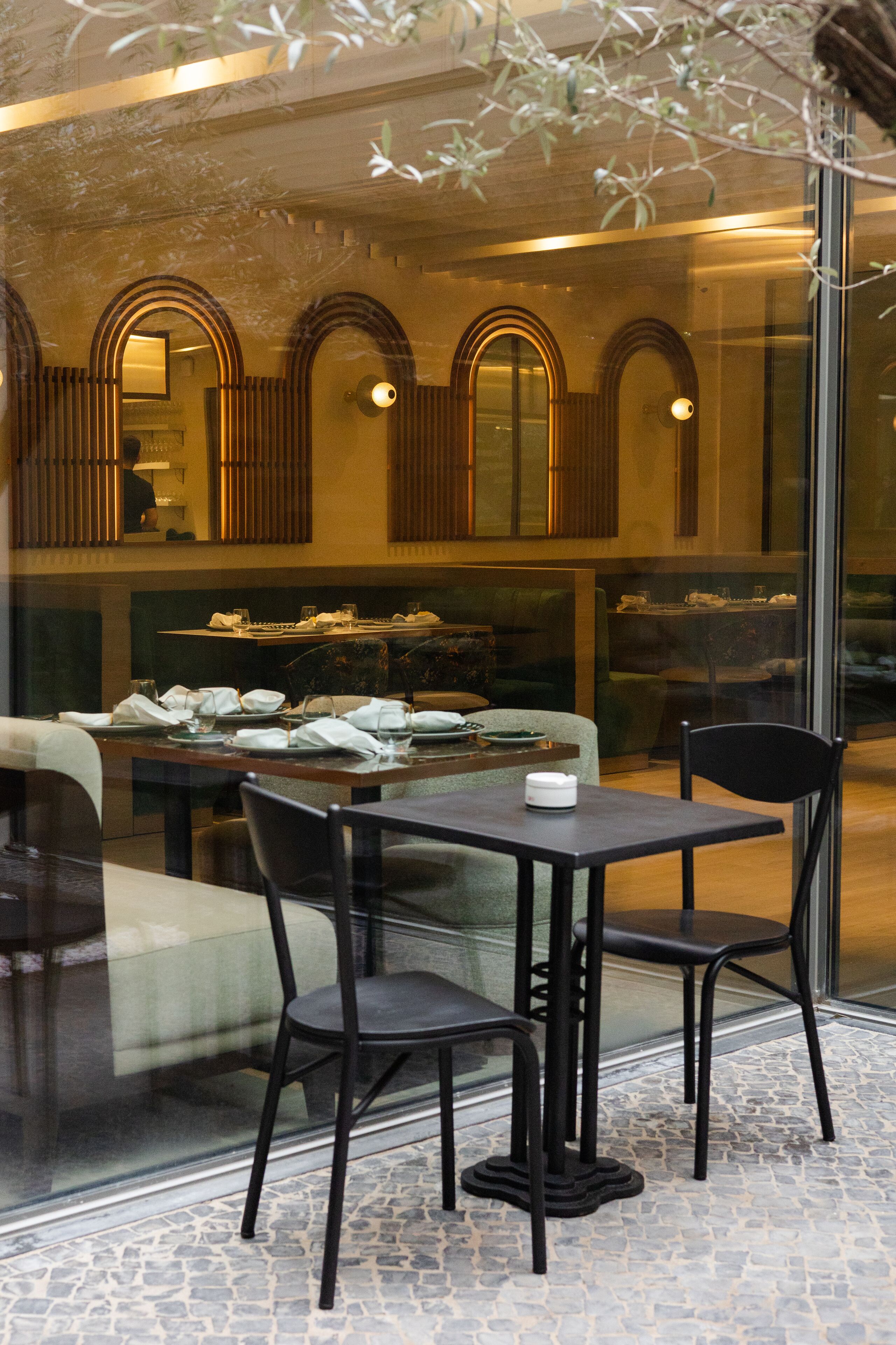 Un restaurante con un separador de vidrio, mesas dispuestas con platos y vasos, y vista de la calle a través del cristal.