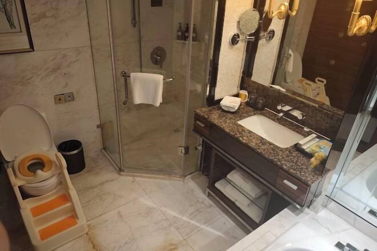Een badkamer met een glazen douchecabine, een toilet met een verhoogd zitje, een wastafel met een marmeren blad en een spiegel met een handdoek aan de muur.