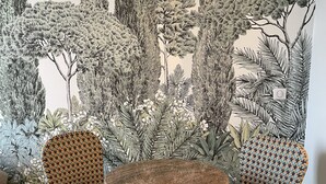 Zona pranzo con due sedie in vimini e un tavolo rotondo, con una parete decorata da un'elegante carta da parati botanica.