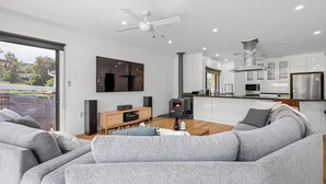 Un salon moderne avec un canapé gris, une table basse en bois et un téléviseur fixé au mur. On y trouve également un coin cuisine avec des meubles blancs et un réfrigérateur en acier inoxydable.