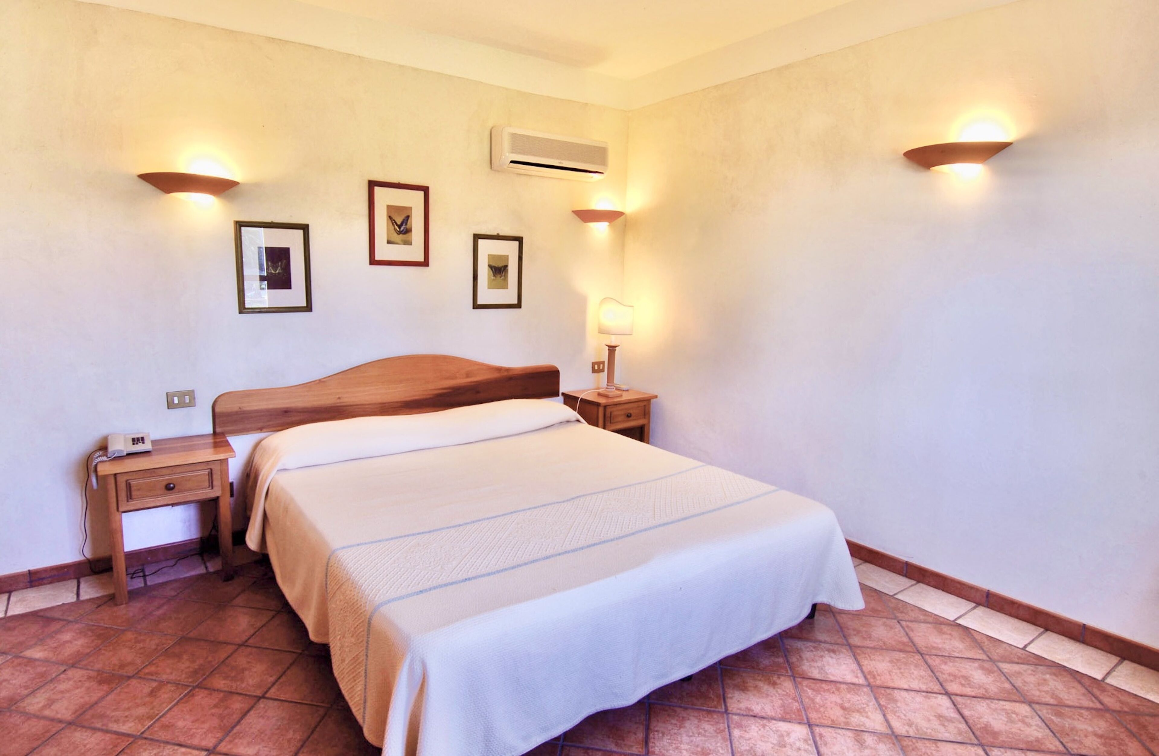 A hotel room with a large bed, a nightstand, a wall-mounted air conditioner, and framed pictures on the wall.