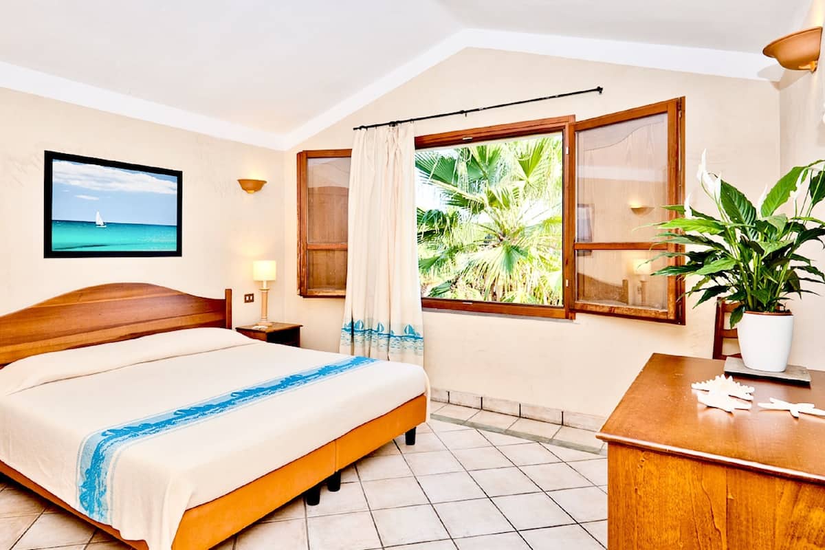 A hotel room with a bed, a desk, a chair, a potted plant, a window with curtains, a wall-mounted TV, and a wall-mounted lamp.