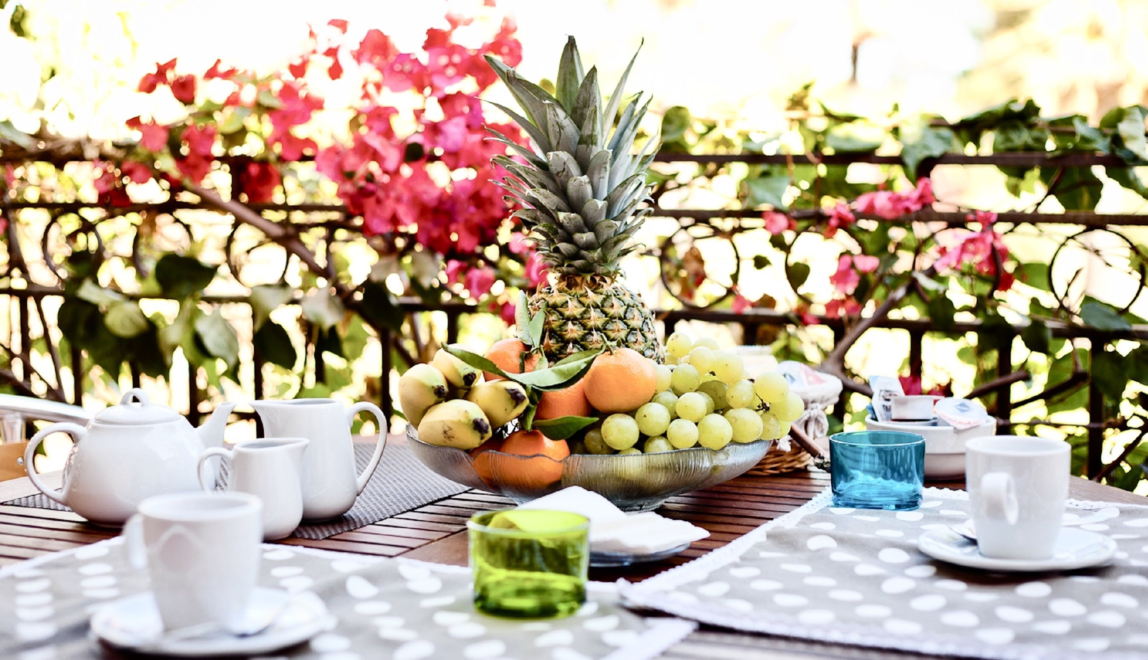 A table set with a pineapple, oranges, grapes, and a bowl of fruit, surrounded by white cups and saucers on a wooden table outdoors.