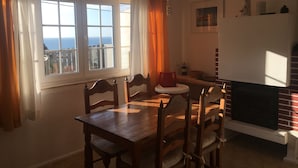 A dining area with a wooden table and chairs, a fireplace, a window with curtains, and a view of the sea.