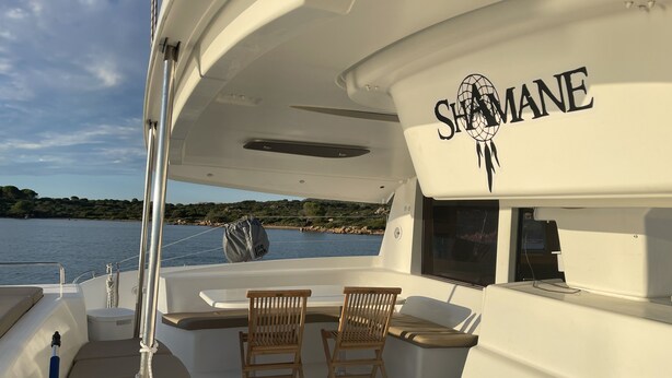 A boat deck with a table and chairs, a 'SHAMANE' logo, and a view of the water and land.