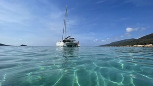 A sailboat on clear turquoise water with a rocky coastline and hills in the background.