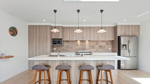 Cocina moderna con isla central, taburetes de madera, iluminación colgante y electrodomésticos de acero inoxidable.