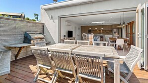 Un patio exterior con mobiliario de madera, una parrilla y vista a una cocina moderna.