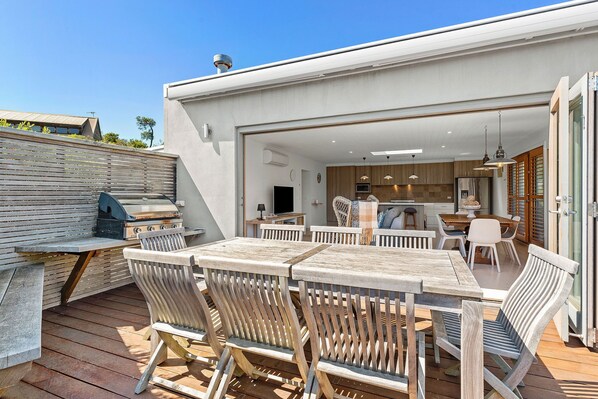 An outdoor patio with wooden furniture, a grill, and a view into a modern kitchen.