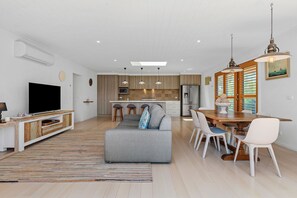 A modern living room with a grey sofa, a flat-screen TV, and a wooden dining table with white chairs.