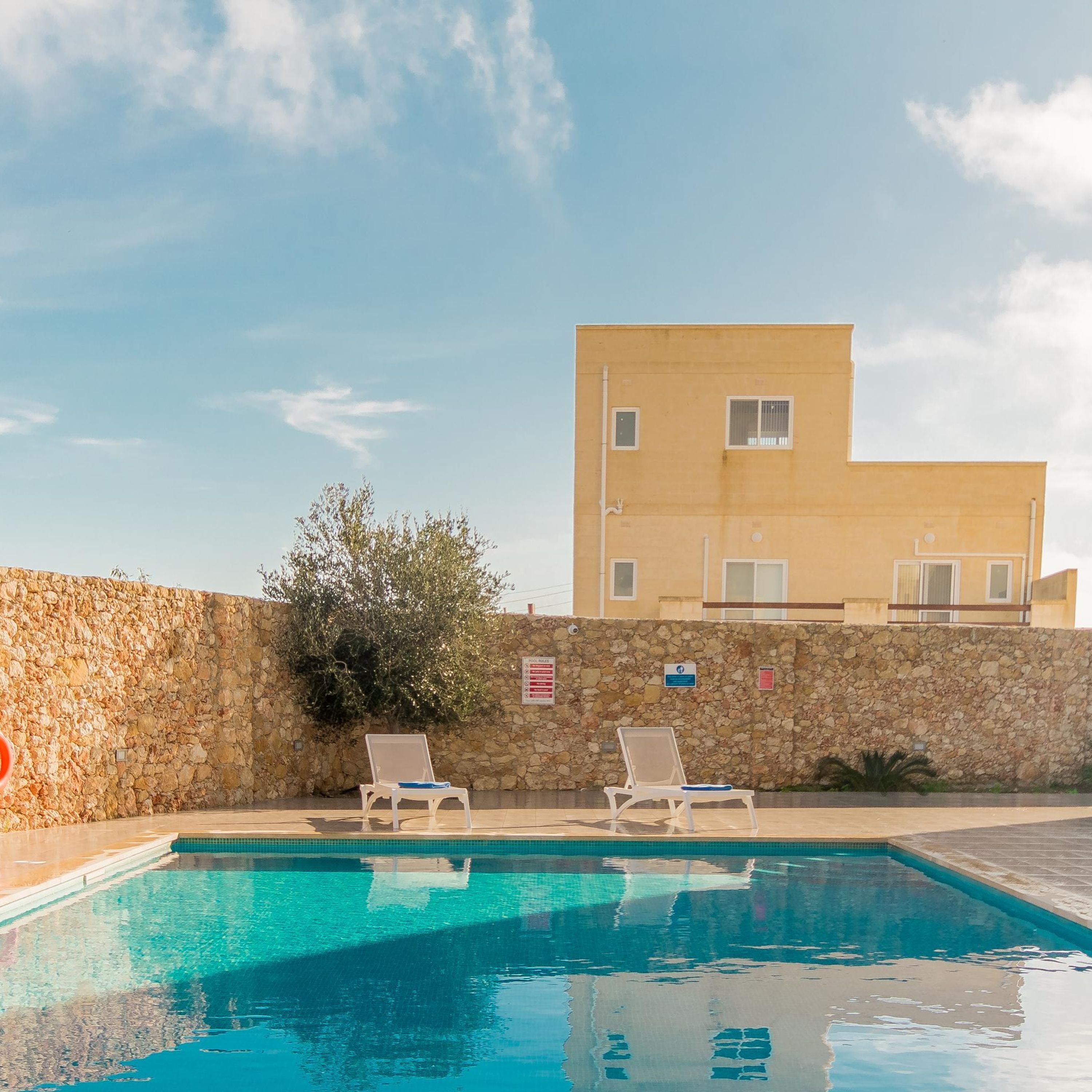 A swimming pool with two lounge chairs, a stone wall, and a building in the background.