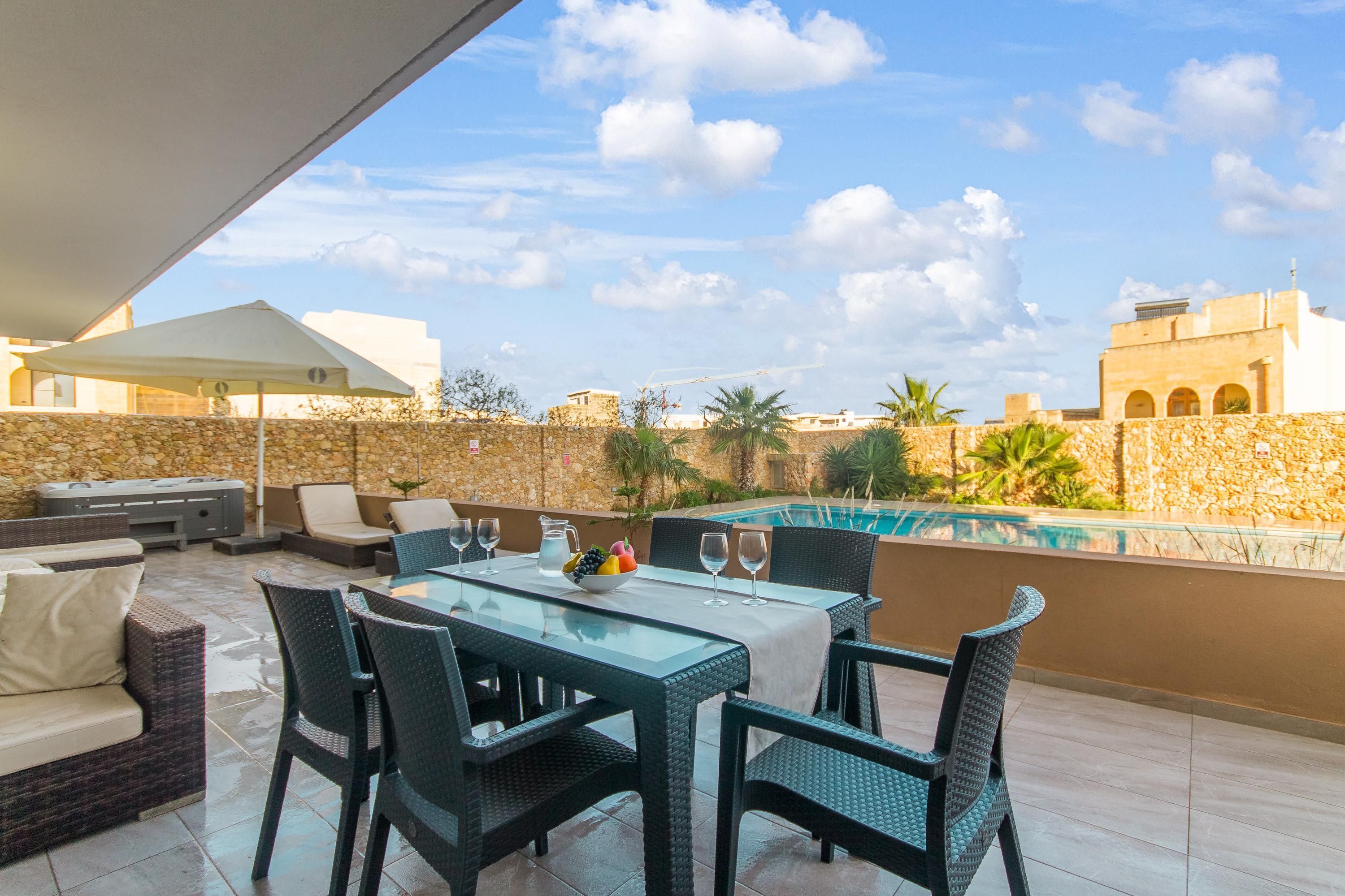 An outdoor patio with a dining table, chairs, and a pool in the background.