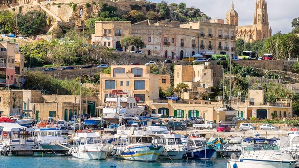 Un pueblo costero con edificios históricos, un puerto deportivo con barcos y una iglesia destacada en una colina.