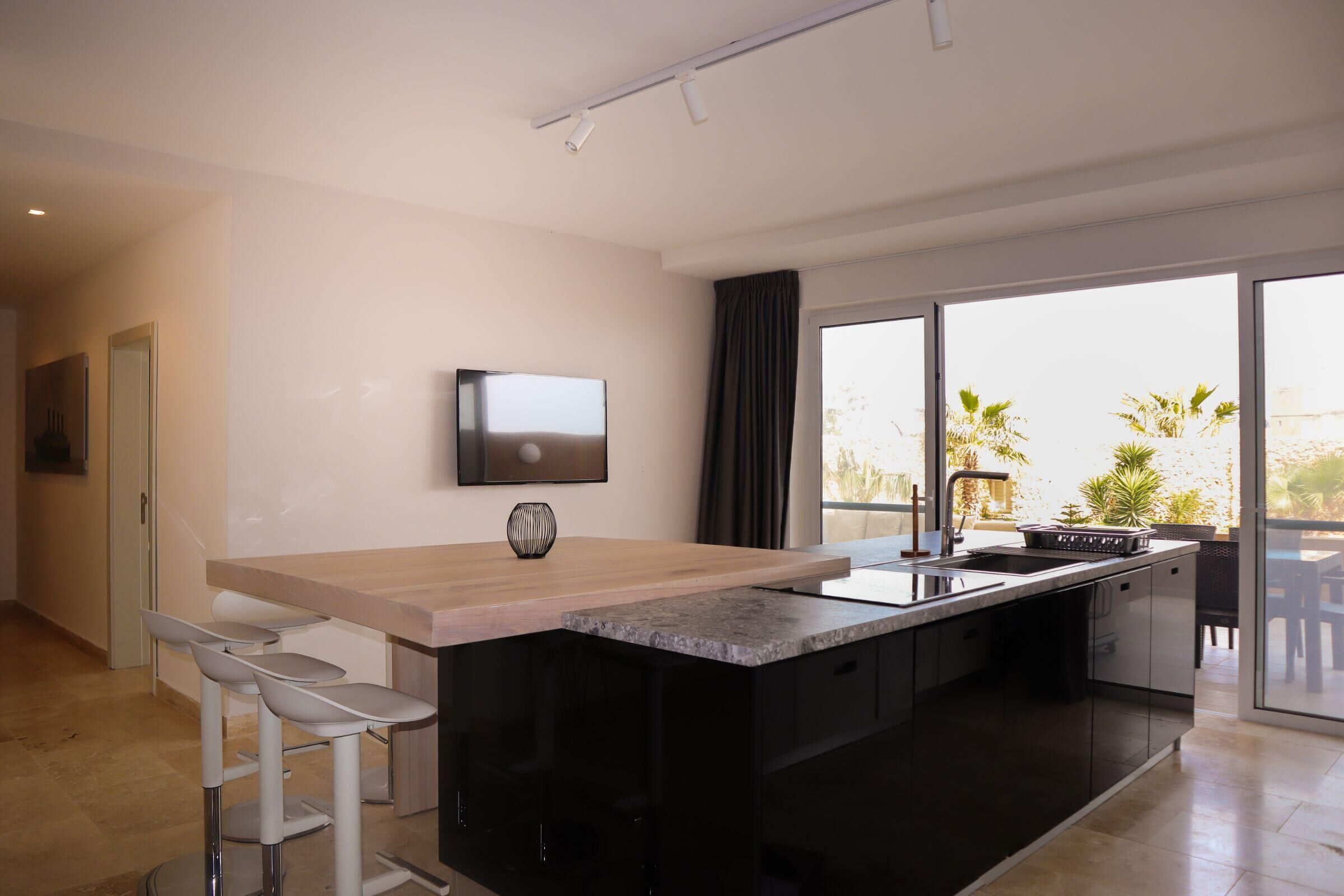 A modern kitchen with a large island, bar stools, a flat-screen TV, and a view of the outdoors.