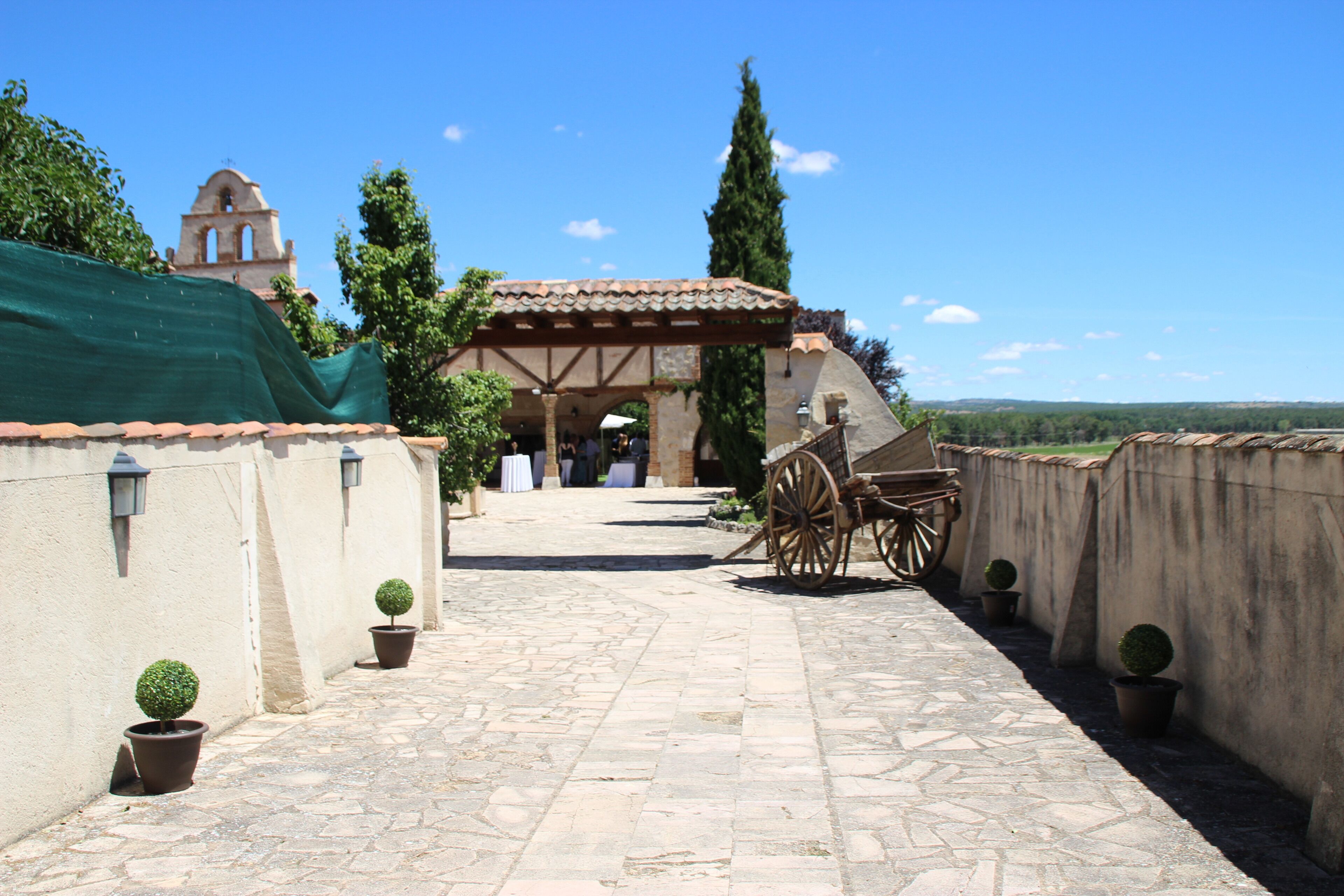 Un patio con un camino de piedra, una zona de estar cubierta y un edificio histórico con una torre de campanas al fondo.