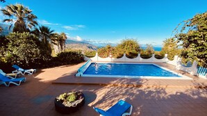 Une piscine avec vue sur les montagnes et la mer, entourée de palmiers et de chaises longues bleues.