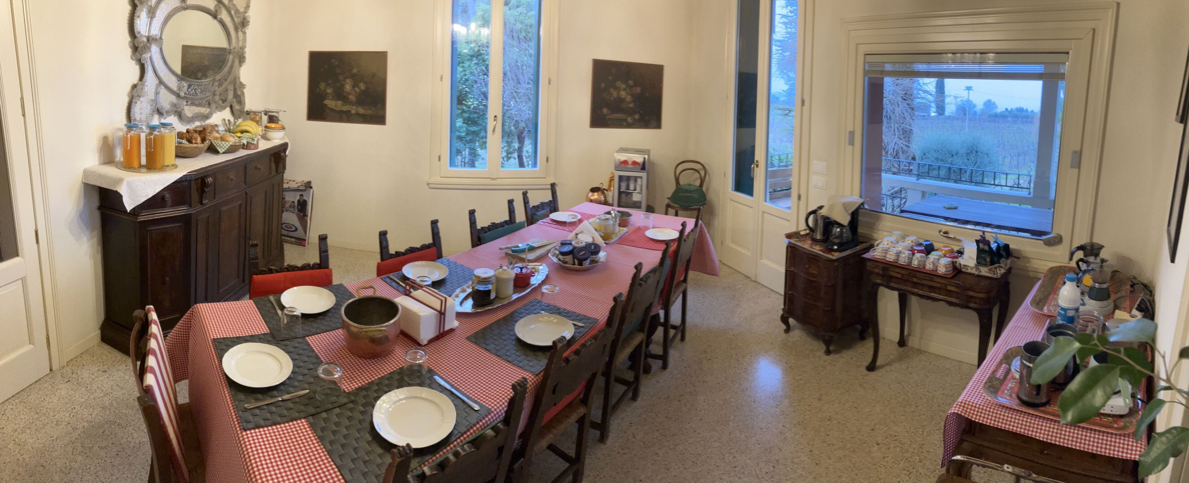 Uma sala de jantar com uma mesa comprida posta para uma refeição, rodeada por cadeiras. Há duas janelas que proporcionam luz natural, e o espaço apresenta uma decoração tradicional com mobiliário de madeira e um espelho na parede.
