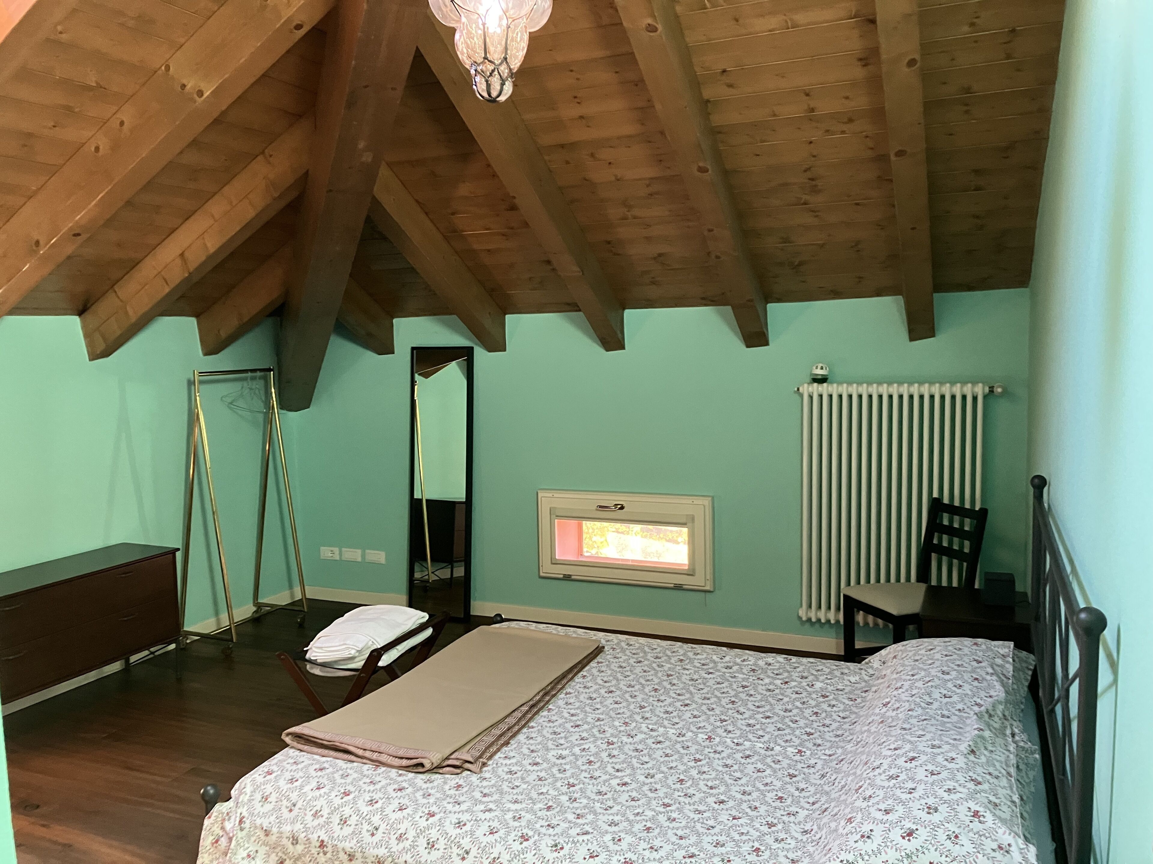 Ein Schlafzimmer mit schrägen Wänden, einem Bett mit floralem Überwurf, einem Nachttisch, einem Sessel, einem Heizkörper und einem Fenster mit Blick.