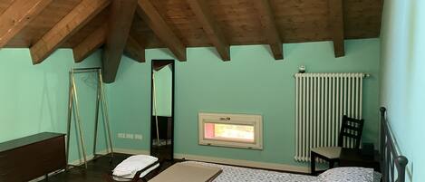 Ein Schlafzimmer mit schrägen Wänden, einem Bett mit floralem Überwurf, einem Nachttisch, einem Sessel, einem Heizkörper und einem Fenster mit Blick.